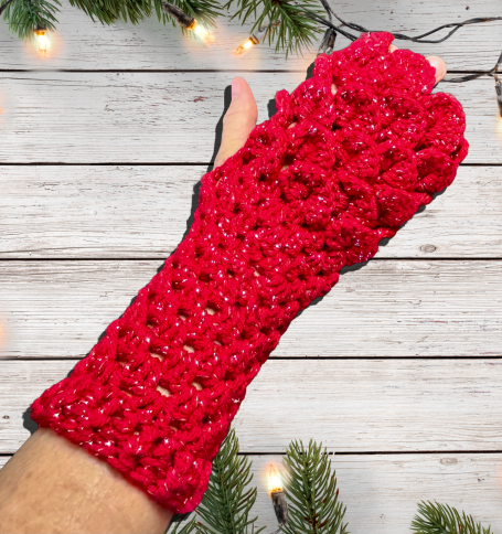 Crocheted Dragon Scale Fingerless Gloves Red Sparkle on a white board background with white Christmas lights and evergreens 